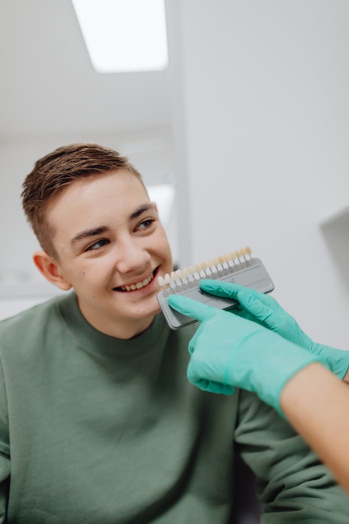 Smiling teenage boy at dental checkup with shade guide in hands of dentist wearing green gloves.