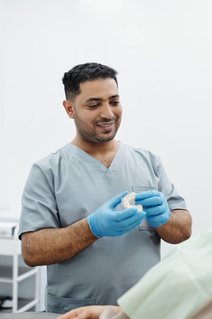 Dentist in scrubs holds dental model and wears blue gloves in a clinic setting.